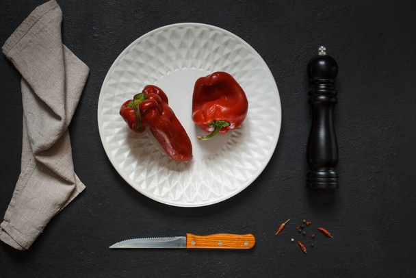 Due brutti peperoni rossi su un piatto bianco, un macinapepe, un tovagliolo di lino e un coltello, su uno sfondo nero, in alto - Foto, immagini