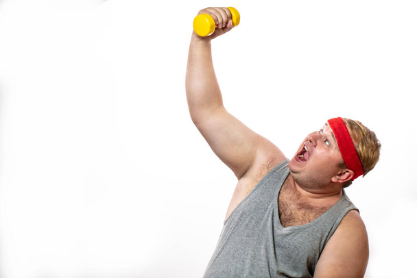 Fat funny man in red headband shows his muscles with dummbell and emotions - Fotografie, Obrázek