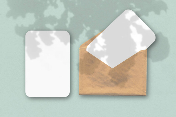 Ein Umschlag mit zwei Blättern strukturierten weißen Papiers auf blauem Tischhintergrund. Mockup mit einer Überlagerung von Pflanzenschatten. Natürliches Licht wirft Schatten von den Eichenblättern - Foto, Bild