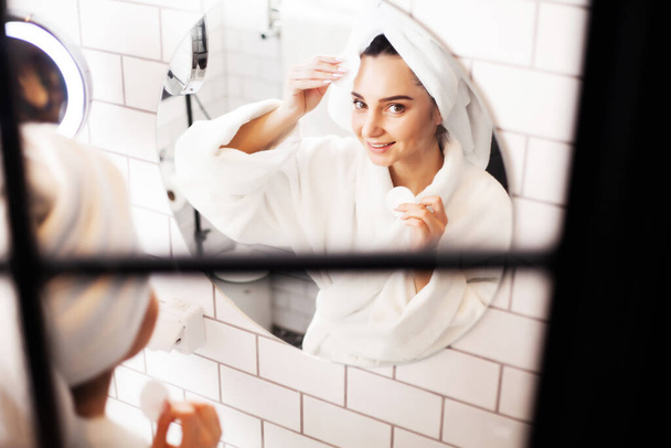 Улыбающаяся молодая женщина умывается губкой для очищения лица в ванной - Фото, изображение