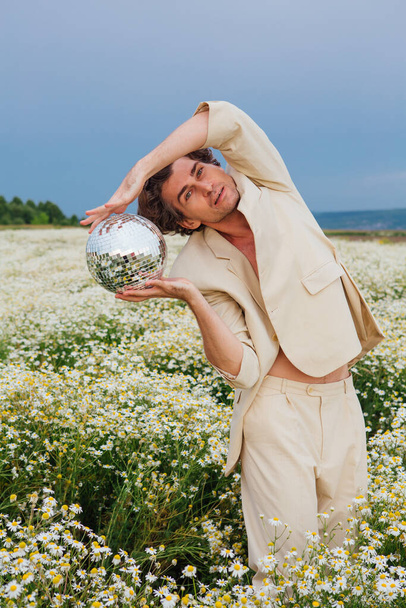 Tall handsome man dressed in a white suit on naked body standing in camomile flowers field, holding mirror disco ball. - Foto, imagen