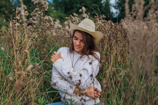 Young beautiful brunette woman dressed in a white sweater, jeans and cowboy straw hat sitting in dry brown bur grass, smiling and laughing during sunset - Foto, immagini