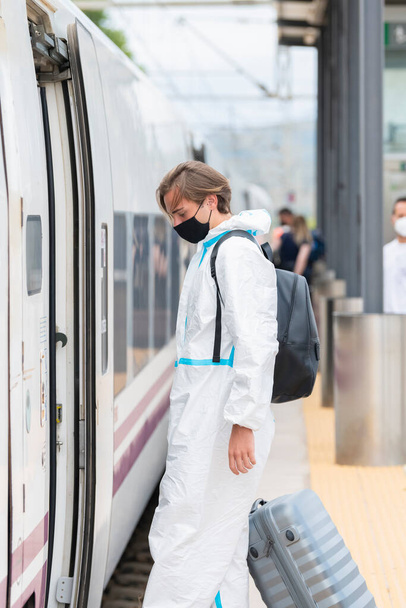 Giovane uomo che indossa una maschera facciale e una tuta protettiva con una valigia che sta per salire su un treno, pur mantenendo una distanza di sicurezza. Turismo e sicurezza. - Foto, immagini