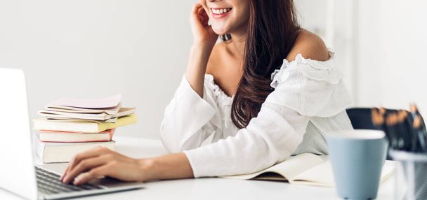Portret uśmiechniętej szczęśliwej pięknej azjatyckiej kobiety relaksującej się przy użyciu technologii laptopa siedząc na stole. Młoda kreatywna dziewczyna pracująca i pisząca na klawiaturze w domu. - Zdjęcie, obraz