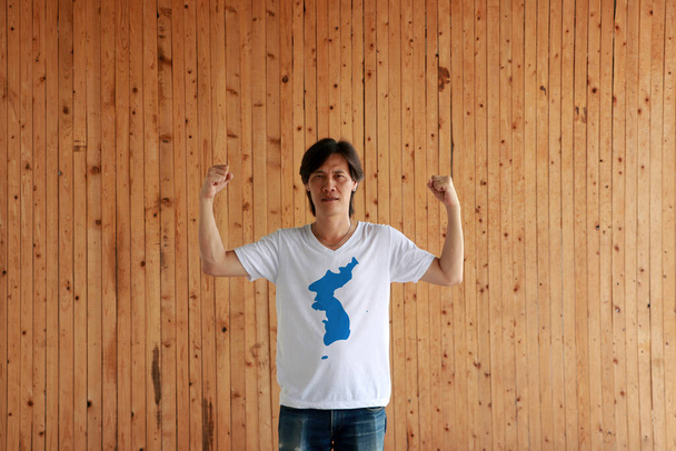 Man met Unification Korea vlag kleur van shirt en staan met opgeheven beide vuisten op de houten muur achtergrond. Koreaans schiereiland in blauw op wit. - Foto, afbeelding