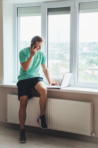 Фото белого человека в спортивной одежде, разговаривающего по телефону и пользующегося ноутбуком у окна - Фото, изображение