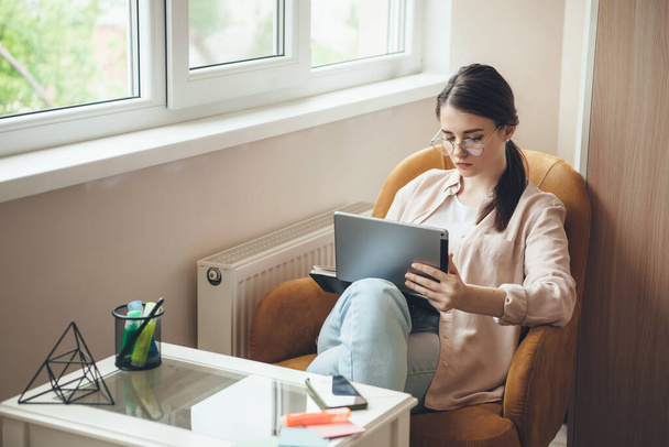 Jovem empresária concentrada com óculos trabalhando no laptop em casa sentada na poltrona
 - Foto, Imagem