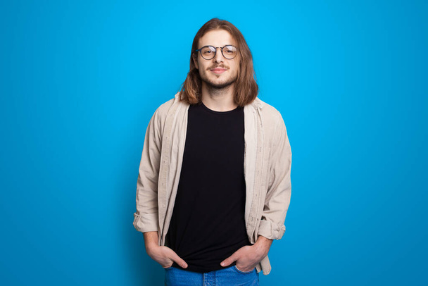 Módní muž s dlouhými vlasy a štětinami pózující s brýlemi na modré zdi studia - Fotografie, Obrázek