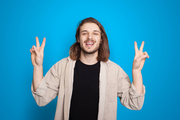 Людина з довгим волоссям, що жестикулює вітання і знак п'єси посміхається на синьому фоні студії - Фото, зображення