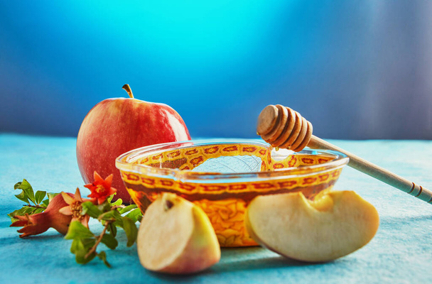 Rosch Haschana ist ein Begriff für das jüdische Neujahrsfest. Honig mit Stäbchen in einer apfelförmigen Schüssel und Granatapfelblättern auf blauem Hintergrund. - Foto, Bild