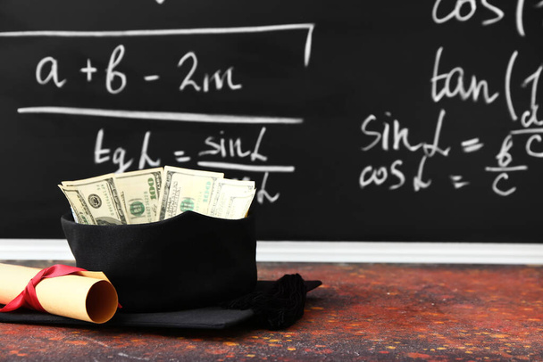 Sombrero de graduación, diploma y dinero en la mesa en el aula. Concepto de tasas - Foto, Imagen