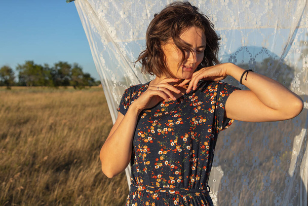 A romantic walk of a curly-haired woman in a dress against the background of the a rack with white curtains, field. The concept of female freedom, emancipation and love - Photo, Image