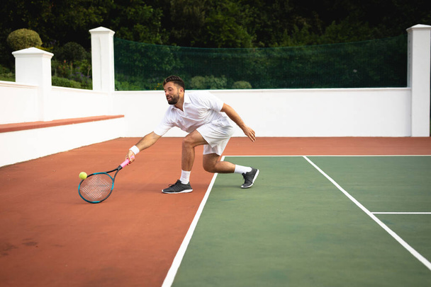 A mixed race man wearing tennis whites spending time on a court playing tennis on a sunny day, hitting a ball with a tennis racket - Photo, Image