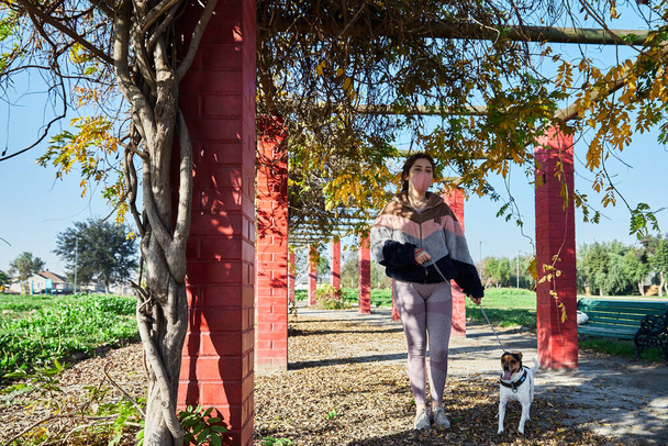 giovane donna che indossa una maschera nel parco a spasso il suo cane in modo sicuro nel bel mezzo della covid 19 pandemia, concetto amante del cane - Foto, immagini