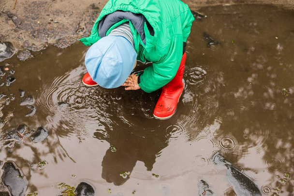 Child. A little boy in a green jacket and red boots plays in the swamp.Carefree childhood. Happy child concept. Happy childhood.He screams and laughs, has fun outside in the swamp. - Foto, afbeelding