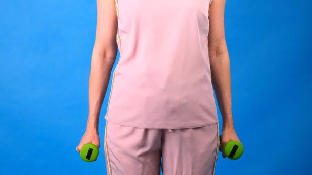 Eine Frau im rosafarbenen Sportanzug mit Hanteln in den Händen macht Übungen auf blauem Hintergrund. Das Konzept von Sport, Ernährung und Gewichtsverlust. - Foto, Bild