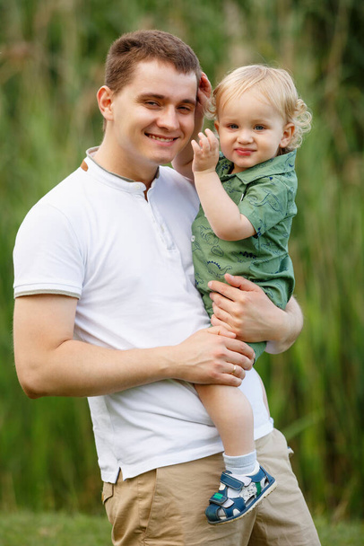 Der Vater hält seinen kleinen Sohn auf dem Arm. Schilf im Hintergrund. Spaziergang im Park. Vertikal gerahmte Aufnahme. - Foto, Bild