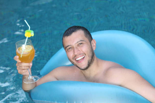 close-up portret van knappe jongeman met cocktail en opblaasbare buis ontspannen in het zwembad - Foto, afbeelding