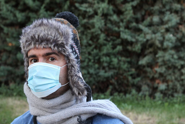 close-up portret van knappe jongeman met masker voor gemoedelijke bescherming en warme kleding in groen park - Foto, afbeelding
