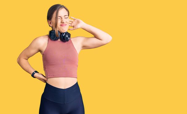 Beautiful caucasian young woman wearing gym clothes and using headphones suffering of neck ache injury, touching neck with hand, muscular pain  - Foto, afbeelding