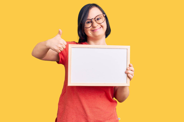 Brunette vrouw met down syndroom met lege witte schoolbord glimlachen blij en positief, duim omhoog doen uitstekend en goedkeuring teken  - Foto, afbeelding