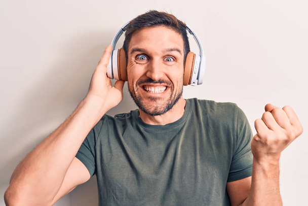 Young handsome man listening to music using headphones over isolated white background screaming proud, celebrating victory and success very excited with raised arm - Foto, Imagen