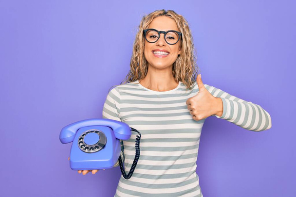Молодая красивая блондинка держит винтажный телефон на изолированном фиолетовом фоне улыбаясь счастливый и позитивный, большой палец вверх делает отлично и знак одобрения - Фото, изображение