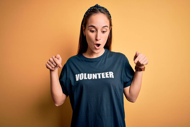 Nuori kaunis nainen yllään vapaaehtoinen t-paita tekee vapaaehtoistyötä keltaisella taustalla Osoittaa alas sormilla näyttää mainosta, yllättynyt kasvot ja avoin suu - Valokuva, kuva