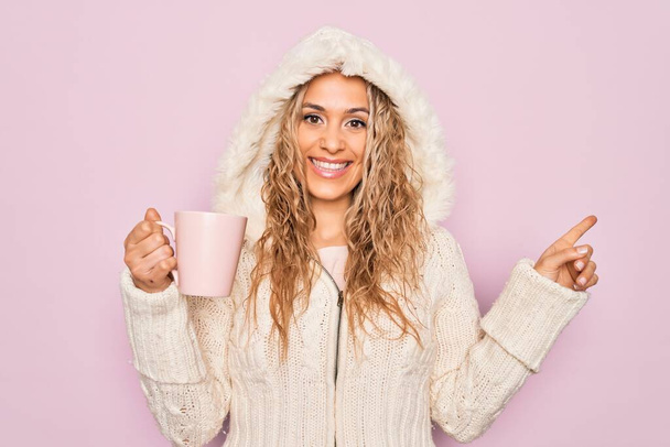 Nuori kaunis blondi nainen yllään rento pusero huppu juominen kuppi kahvia Hymyilevä onnellinen osoittaa kädellä ja sormella sivulle - Valokuva, kuva