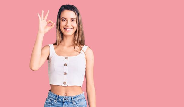 Young beautiful girl wearing casual sleeveless t shirt smiling positive doing ok sign with hand and fingers. successful expression.  - Photo, Image