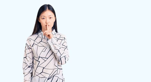 Νεαρή όμορφη Κινέζα γυναίκα που φοράει casual πουκάμισο ζητώντας να είναι ήσυχη με το δάχτυλο στα χείλη. σιωπή και μυστική έννοια.  - Φωτογραφία, εικόνα