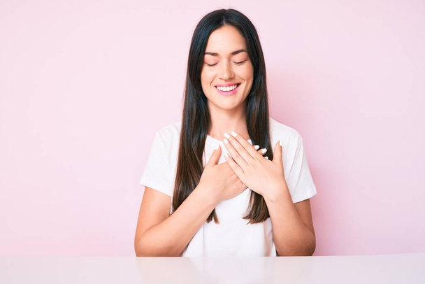 Young caucasian woman sitting at the table wearing casual white tshirt smiling with hands on chest with closed eyes and grateful gesture on face. health concept.  - Photo, image