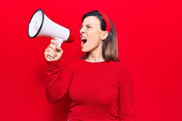 Νεαρή όμορφη γυναίκα που φοράει ντάιαντεμ με θυμωμένη έκφραση. Φωνάζοντας δυνατά χρησιμοποιώντας μεγάφωνο που στέκεται πάνω από απομονωμένο κόκκινο φόντο - Φωτογραφία, εικόνα