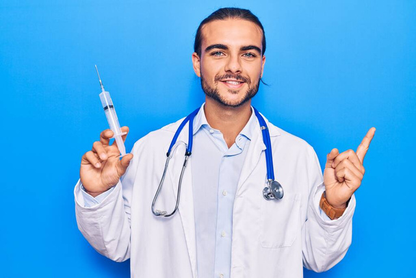 Jovem homem bonito vestindo médico estetoscópio segurando seringa sorrindo feliz apontando com a mão e o dedo para o lado
  - Foto, Imagem