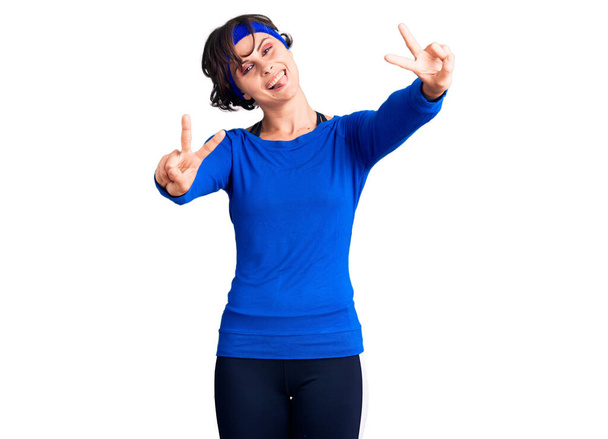 Красивая молодая женщина с короткими волосами в тренировочной одежде, улыбаясь языком, показывает пальцы обеих рук, делающих знак победы. Номер два.  - Фото, изображение