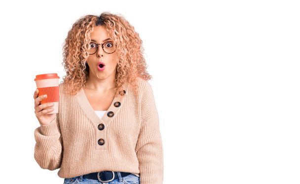 Young blonde woman with curly hair wearing glasses and drinking a cup of coffee scared and amazed with open mouth for surprise, disbelief face  - Photo, Image