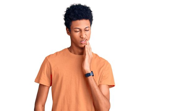 歯の痛みや歯の病気のため、若いアフリカ系アメリカ人男性は痛みを伴う表情で手で口に触れるカジュアルな服を着ています。歯科医  - 写真・画像