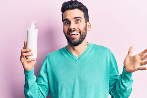 Young handsome man with beard holding bottle of cream celebrating achievement with happy smile and winner expression with raised hand  - Foto, afbeelding