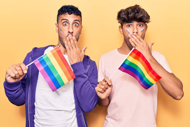 Молодая пара геев с радужными флагами lgbtq закрывает рот рукой, шокирована и боится ошибиться. Удивительное выражение  - Фото, изображение