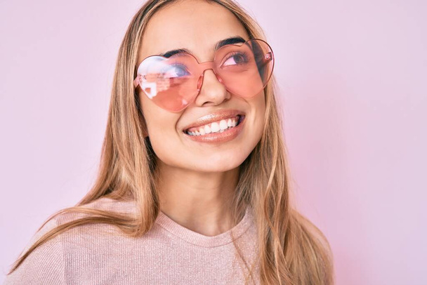 Young beautiful blonde woman wearing heart shaped sunglasses smiling looking to the side and staring away thinking.  - Photo, Image