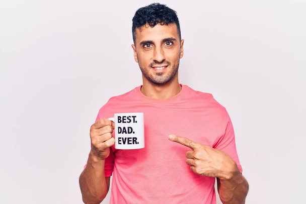 Giovane uomo ispanico bere tazza di caffè con il miglior papà mai messaggio sorridente felice puntando con mano e dito  - Foto, immagini