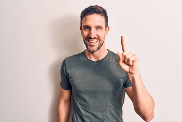 Young handsome man wearing casual t-shirt standing over isolated white background smiling with an idea or question pointing finger up with happy face, number one - Photo, Image