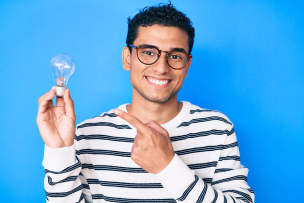 Young handsome hispanic man holding lightbulb for inspiration and idea smiling happy pointing with hand and finger  - Photo, Image