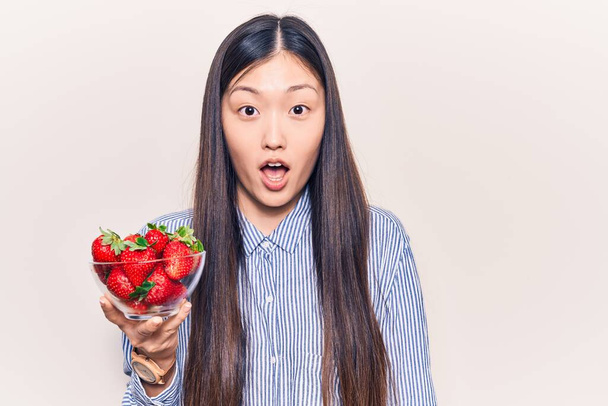 Νεαρή όμορφη Κινέζα γυναίκα κρατώντας μπολ με φράουλες φοβισμένη και κατάπληκτη με ανοιχτό στόμα για έκπληξη, δυσπιστία πρόσωπο  - Φωτογραφία, εικόνα