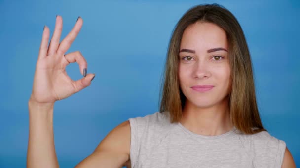 Beautiful woman in gray t-shirt shows ok, winks eye, smiles on blue background - Footage, Video