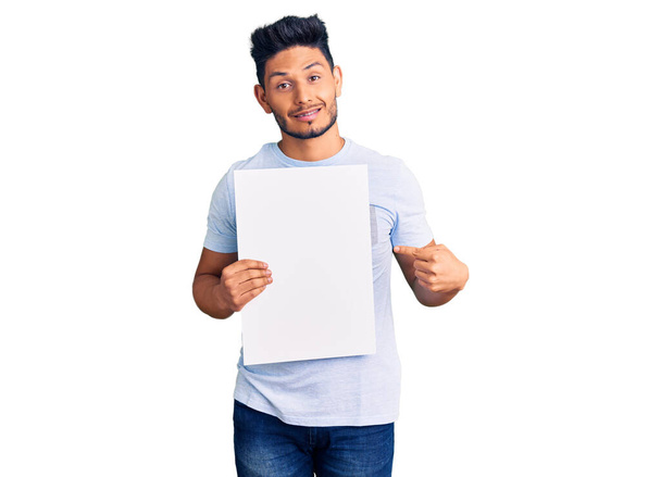 Handsome latin american young man holding cardboard banner with blank space smiling happy pointing with hand and finger  - Foto, Imagen