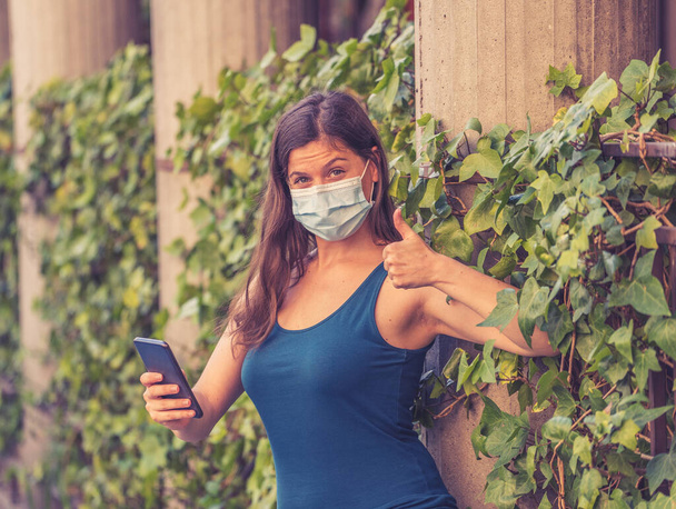 Вспышка COVID-19. Молодая женщина, идущая по городской улице в защитной хирургической маске для лица и пользующаяся мобильным телефоном летом. Коронавирус, новое нормальное и обязательное использование маски для лица - Фото, изображение