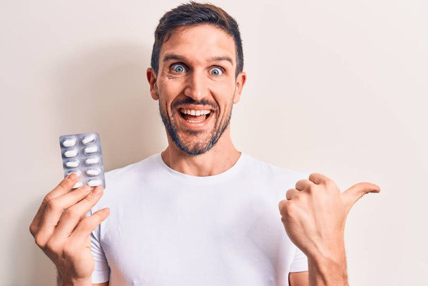 Young handsome man holding medicine pills standing over isolated white background pointing thumb up to the side smiling happy with open mouth - Foto, Imagem