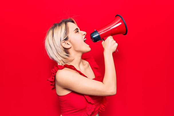 Young beautiful blonde woman with angry expression. Screaming loud using megaphone standing over isolated red background - Fotografie, Obrázek
