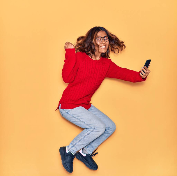 Young beautiful latin woman wearing glasses smiling happy. Jumping with smile on face make selfie by smartphone over isolated yellow background - 写真・画像
